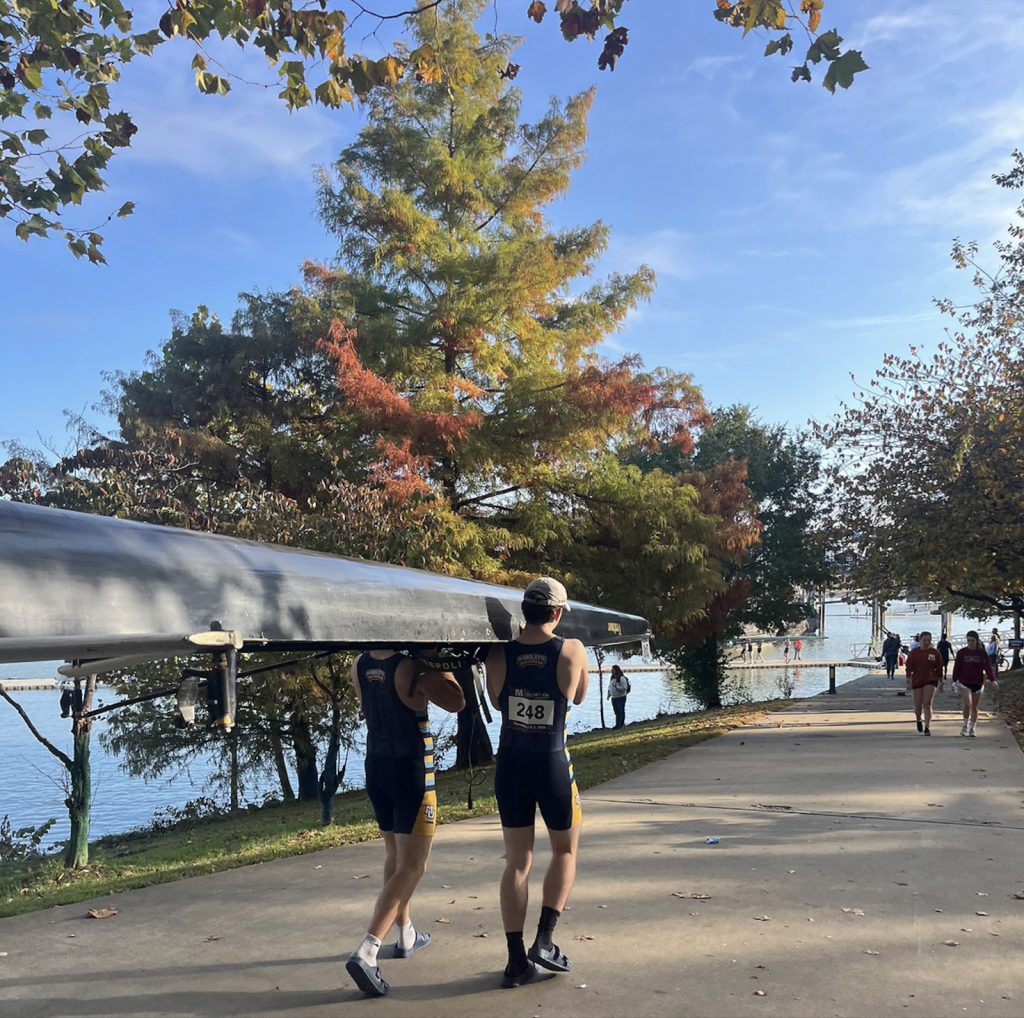 Two Marquette rowers carry a shell to the water under fall color.